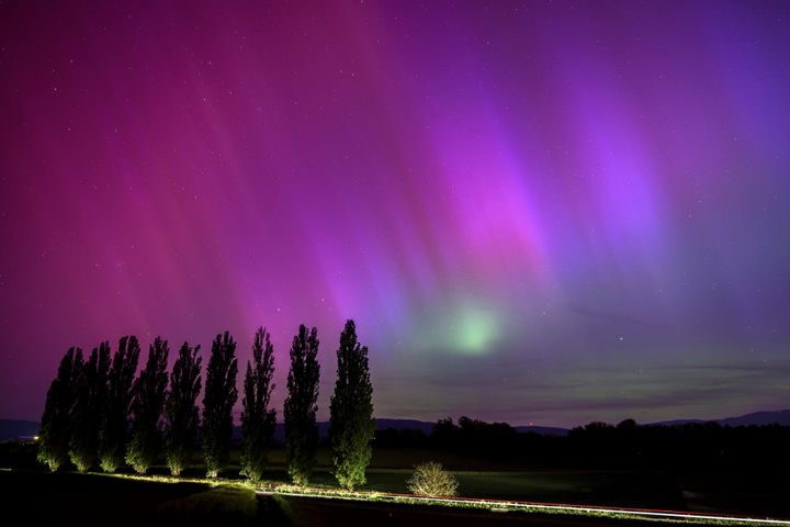 In this long exposure photograph, a car drives past and illuminates poplars as the northern lights glow in the night sky above the village of Daillens, Switzerland, early Saturday, May 11, 2024.