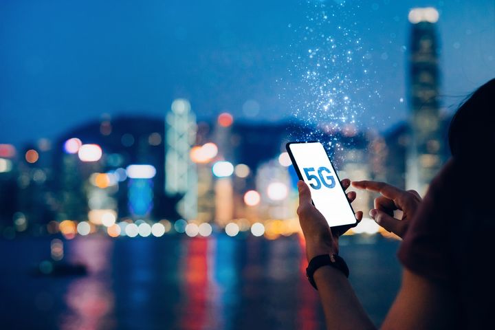 Young woman using a smartphone that says 5G against the iconic city skyline of Hong Kong by the promenade of Victoria Harbour at night, with the concept of 5G communications dissolved into light particles